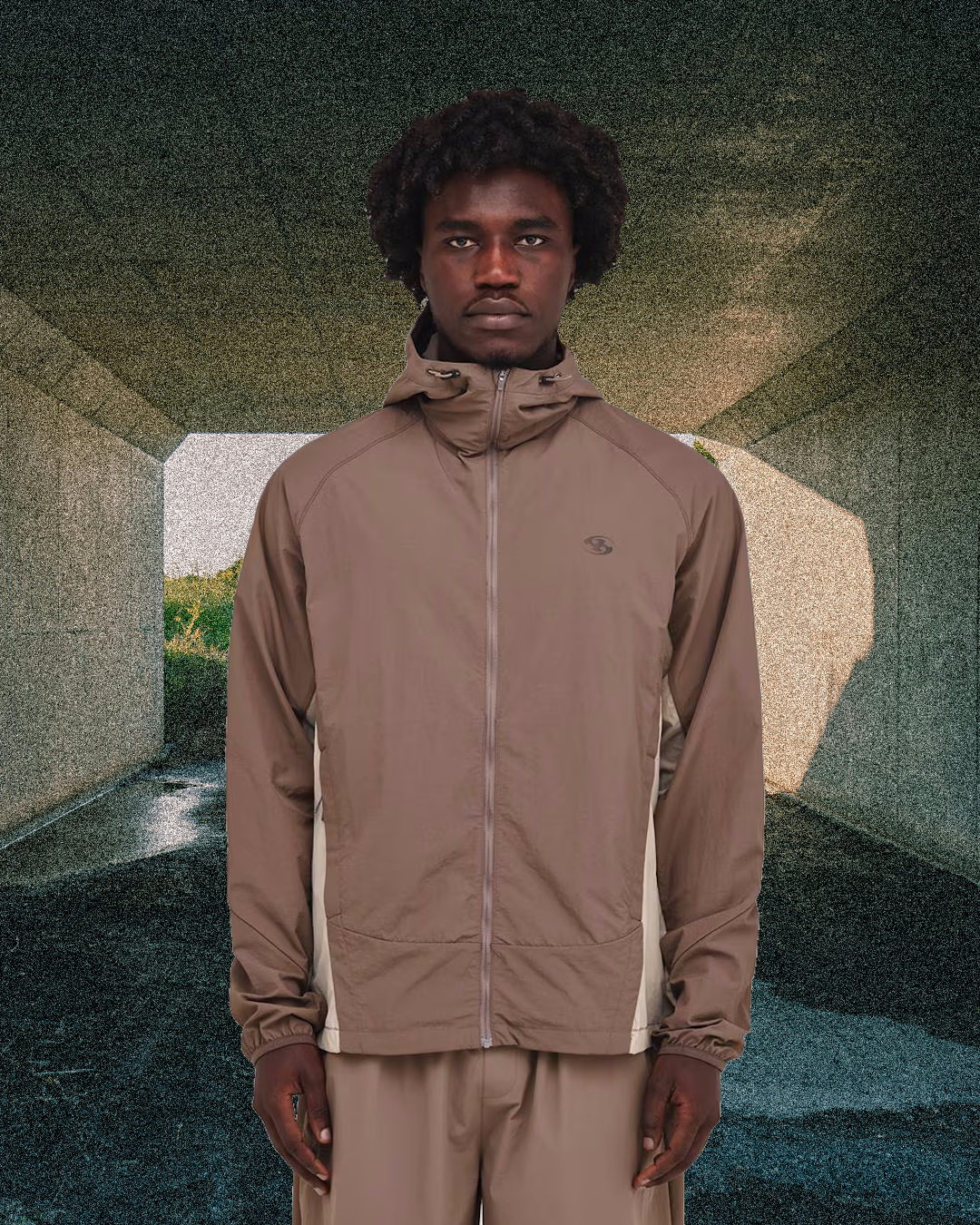 Man in a brown hooded windbreaker with white side panels in a textured tunnel.
