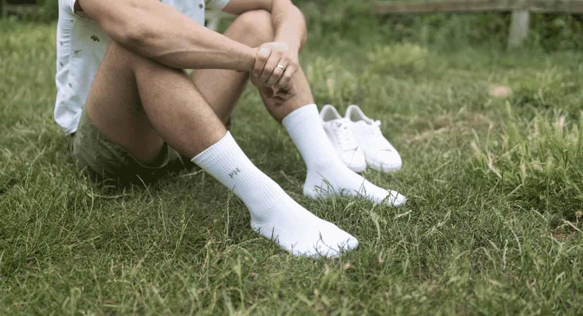 A man in shorts and white ribbed socks sitting on green grass with white sneakers nearby.