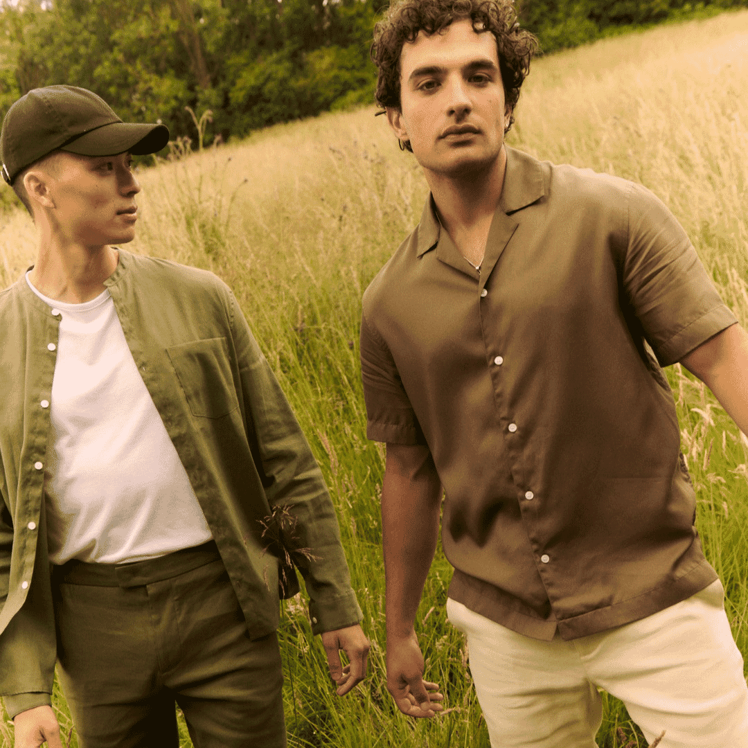 Two men in earth-toned outfits—olive button-up jacket and brown short-sleeve shirt—walking through a sunlit field.