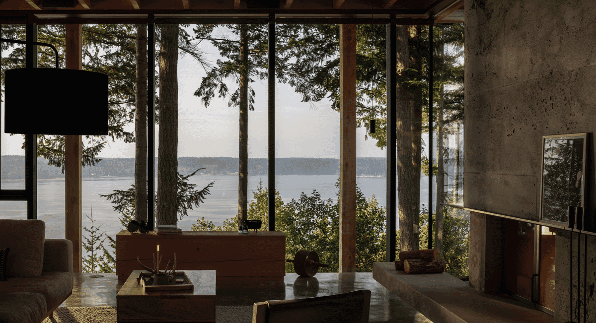 Interior of a minimalist forest cabin with expansive views of the water through large glass windows.