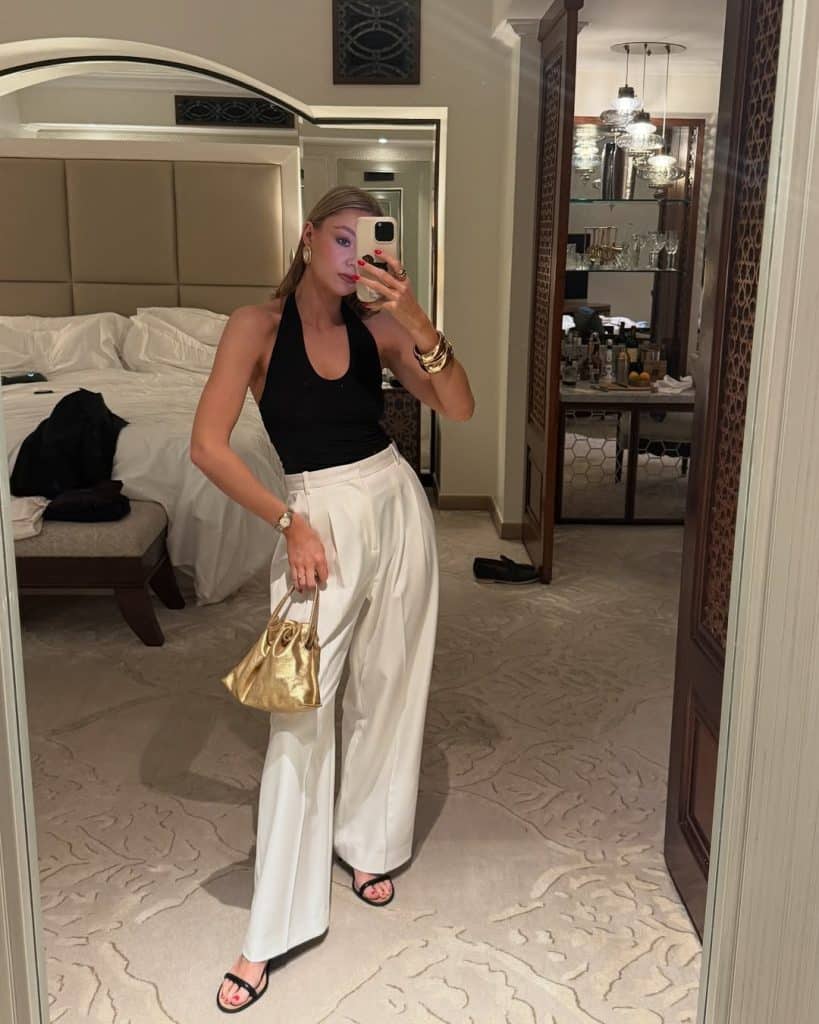 Woman in a black halter top and white wide-leg trousers holding a metallic gold bag.