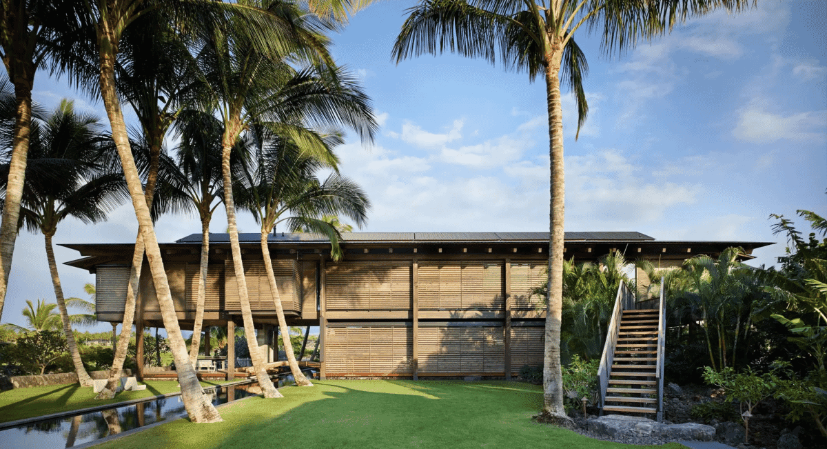The exterior of a tropical residence featuring intricate wooden slat walls and palm trees under a clear sky.