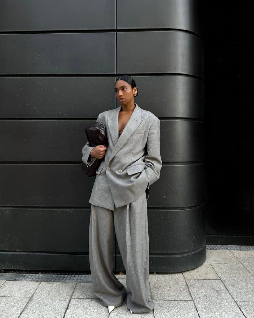 A woman in a full oversized grey wool suit with wide-leg trousers standing against a dark wall.