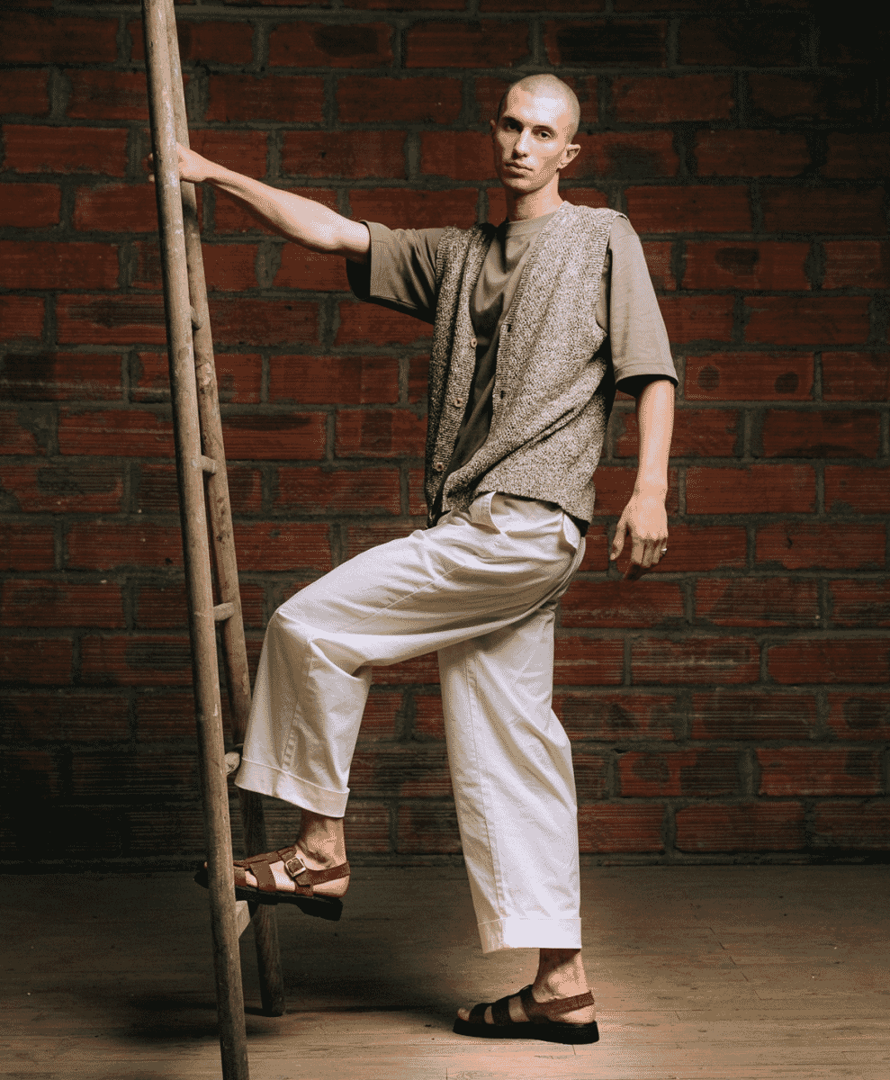 A man in a textured knit vest and wide-leg white pants wearing brown fisherman sandals on a ladder.