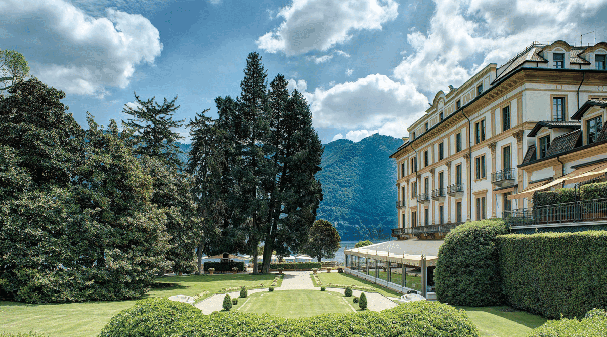 A grand European-style hotel façade with manicured green gardens and a mountain view.