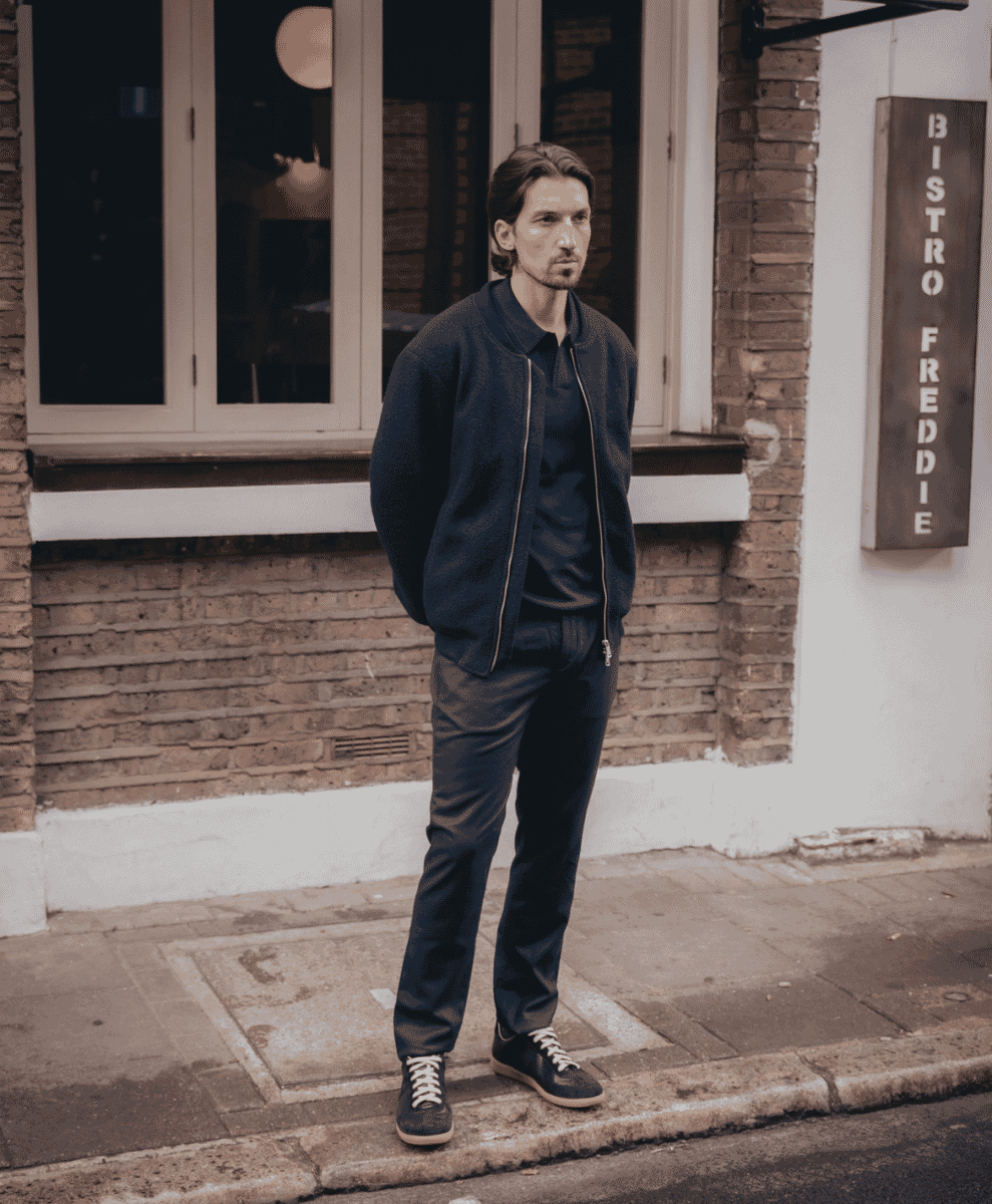 Man in a dark bomber jacket and matching pants standing on a sidewalk near a bistro.