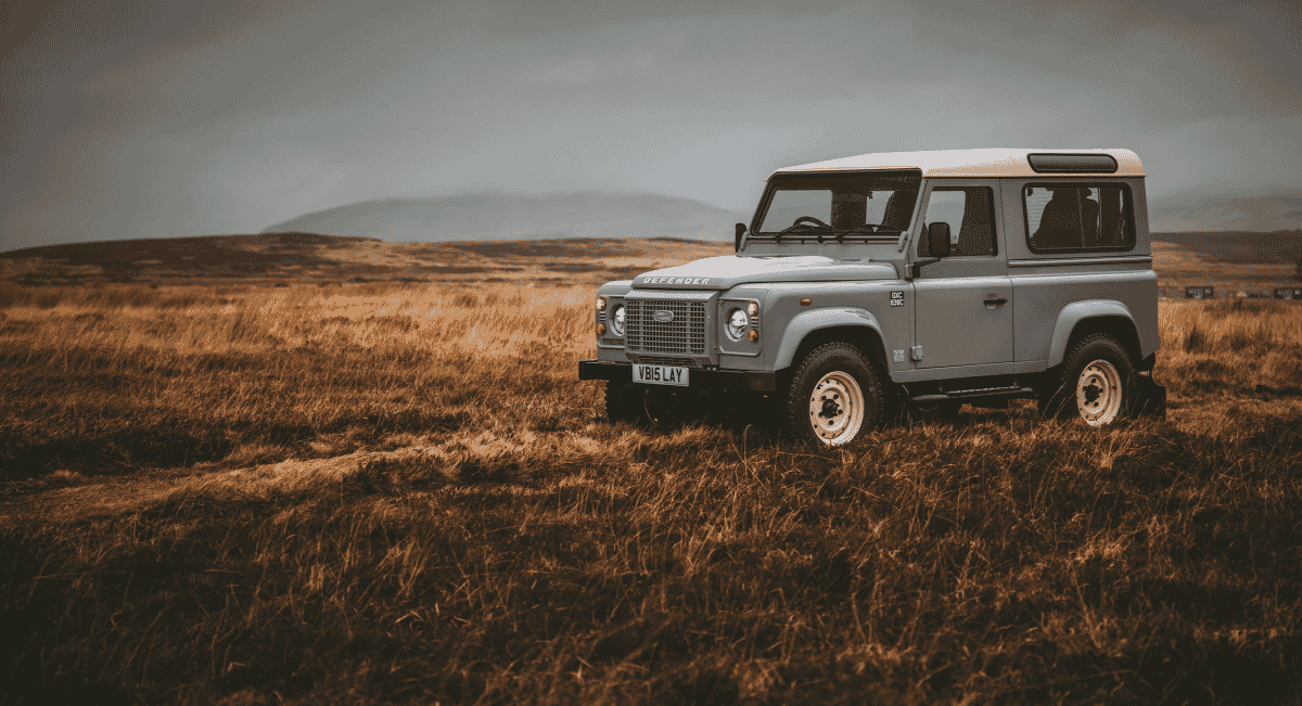 A silver Land Rover Defender standing in a rugged, grassy field under a cloudy sky.