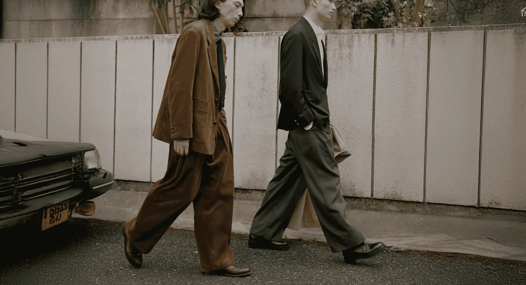 Two men walking together, one in a brown corduroy suit and the other in a dark tailored suit with relaxed pants.