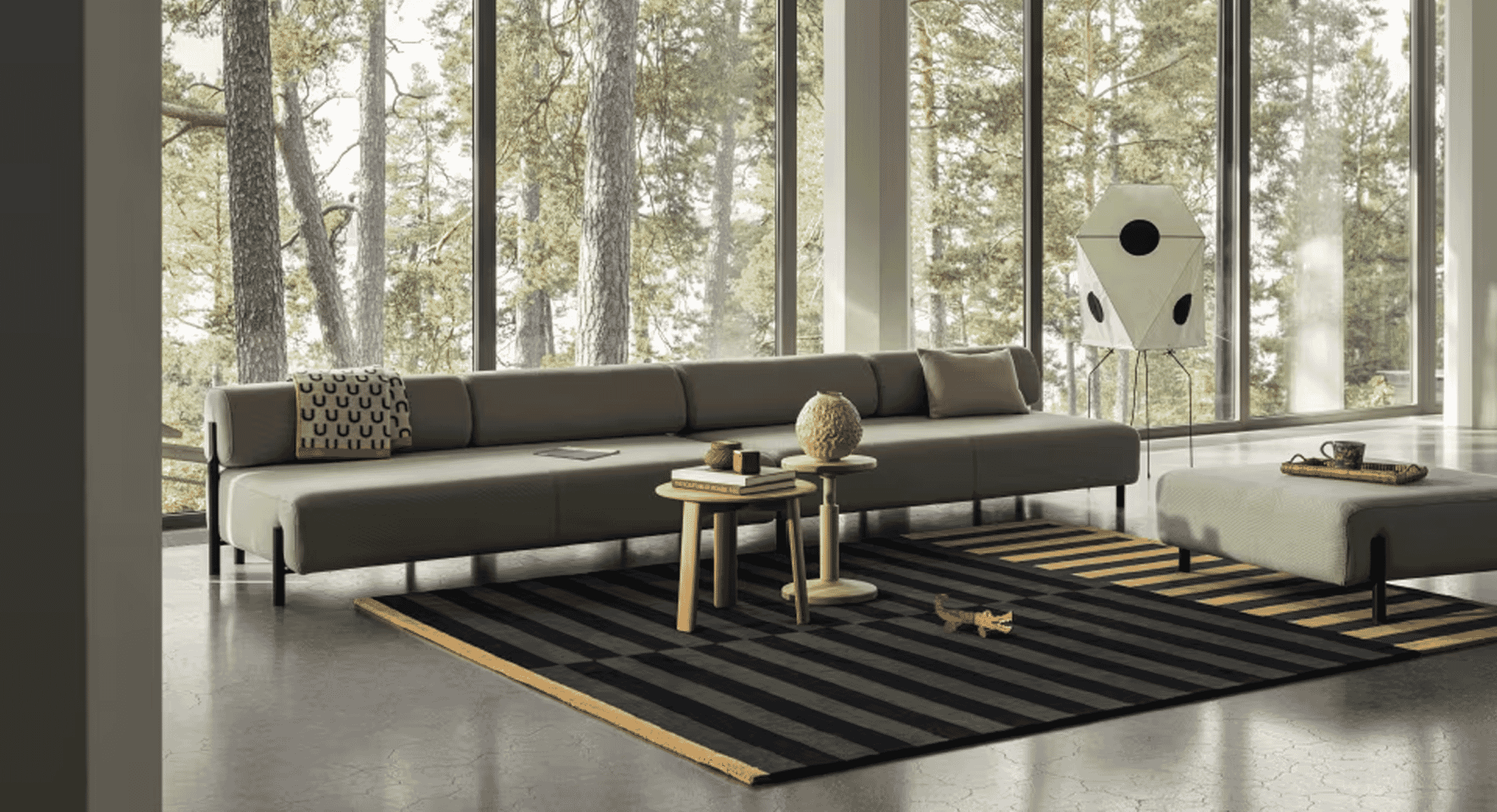 Wide-angle view of a modern living room with a gray sectional sofa, striped rug, and forest background through floor-to-ceiling windows.