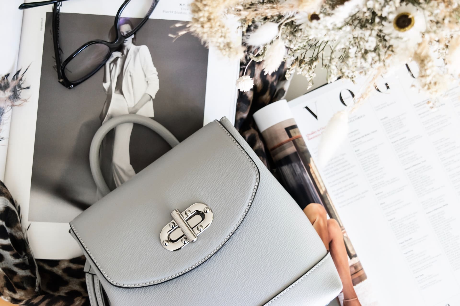 A light gray structured handbag and fashion magazines on a flat-lay with glasses and leopard-print accents.