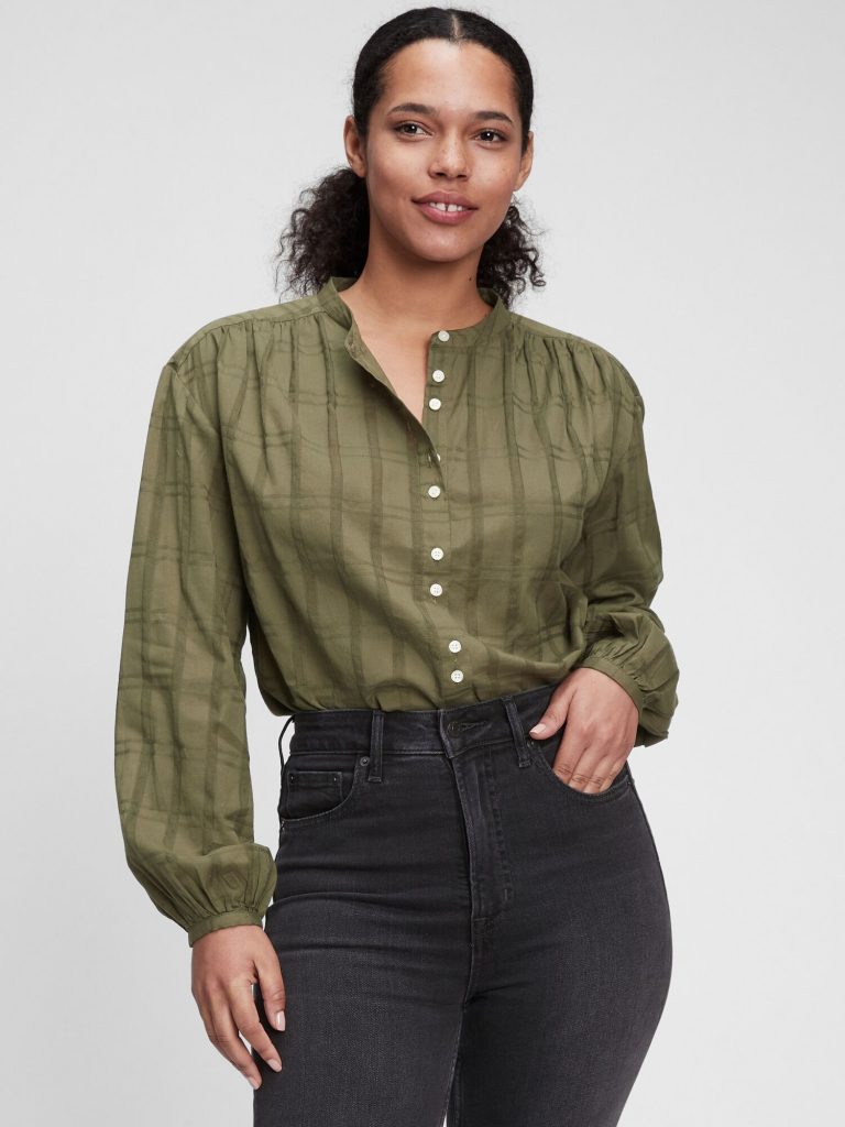 Woman in an olive green plaid blouse with puff sleeves and dark jeans.