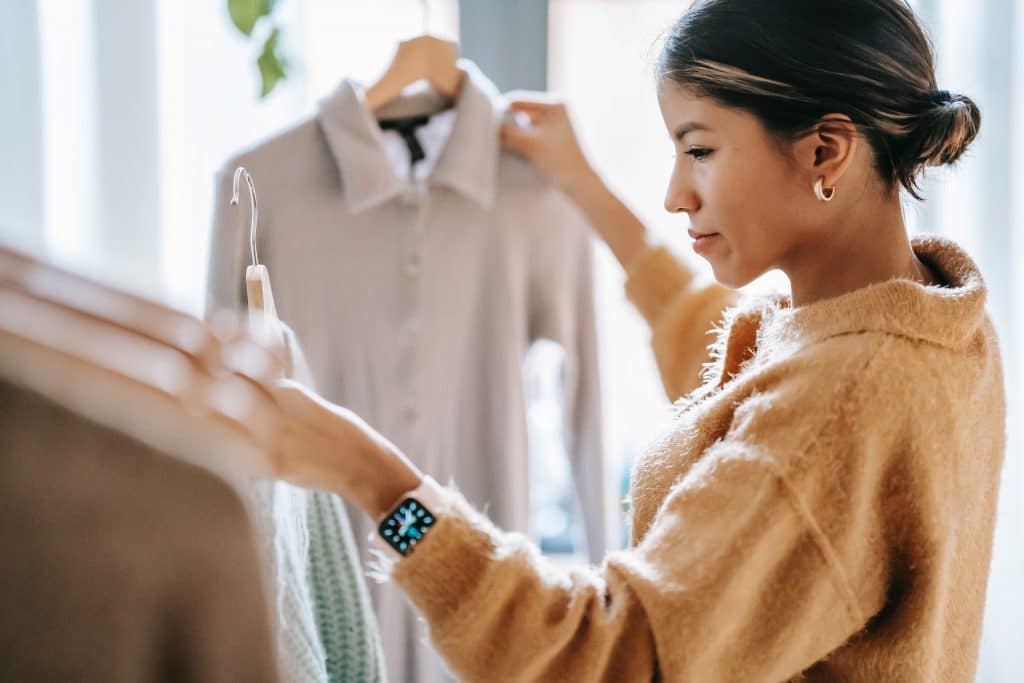 A woman in a beige fuzzy sweater browsing through high-quality clothing on a rack in a sunlit room.