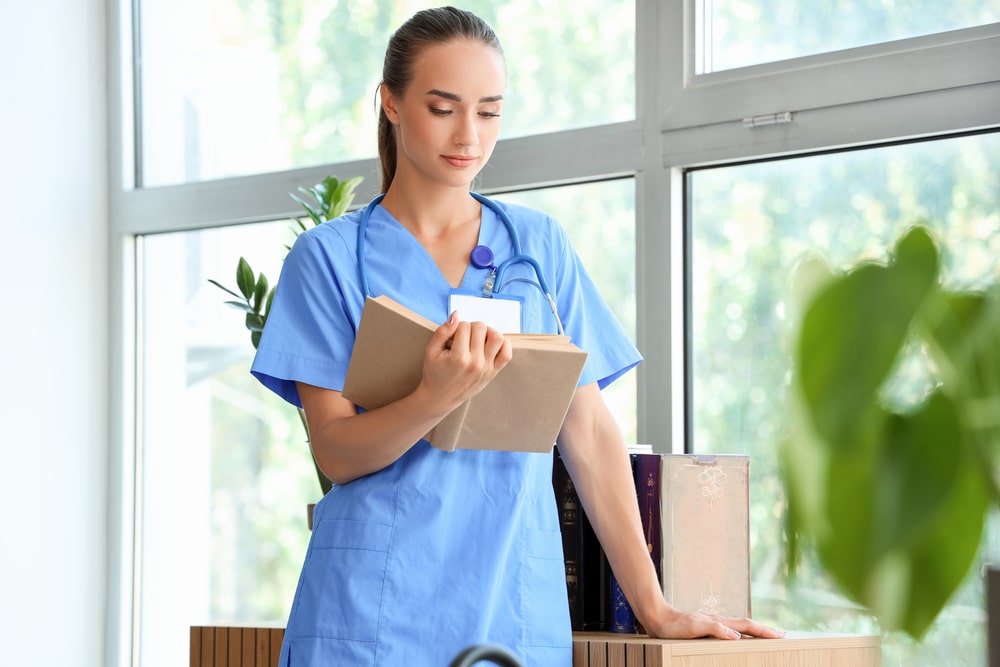 A healthcare professional in light blue scrubs reading a medical book near a window with greenery.
