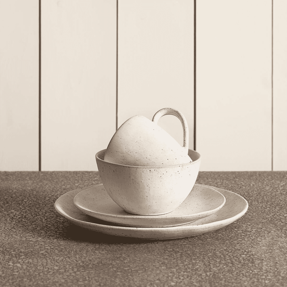 Speckled ceramic cups and plates stacked against a vertical textured panel in sepia tones.