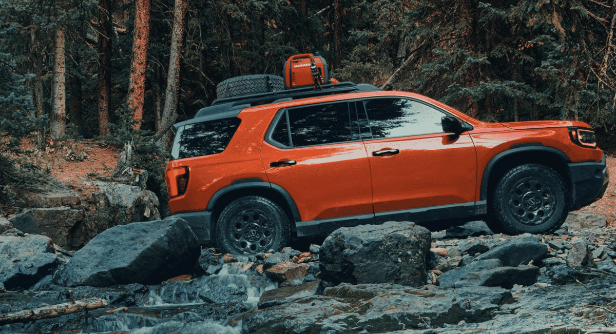 An orange off-road SUV equipped with a roof rack and spare tire on rocky terrain.
