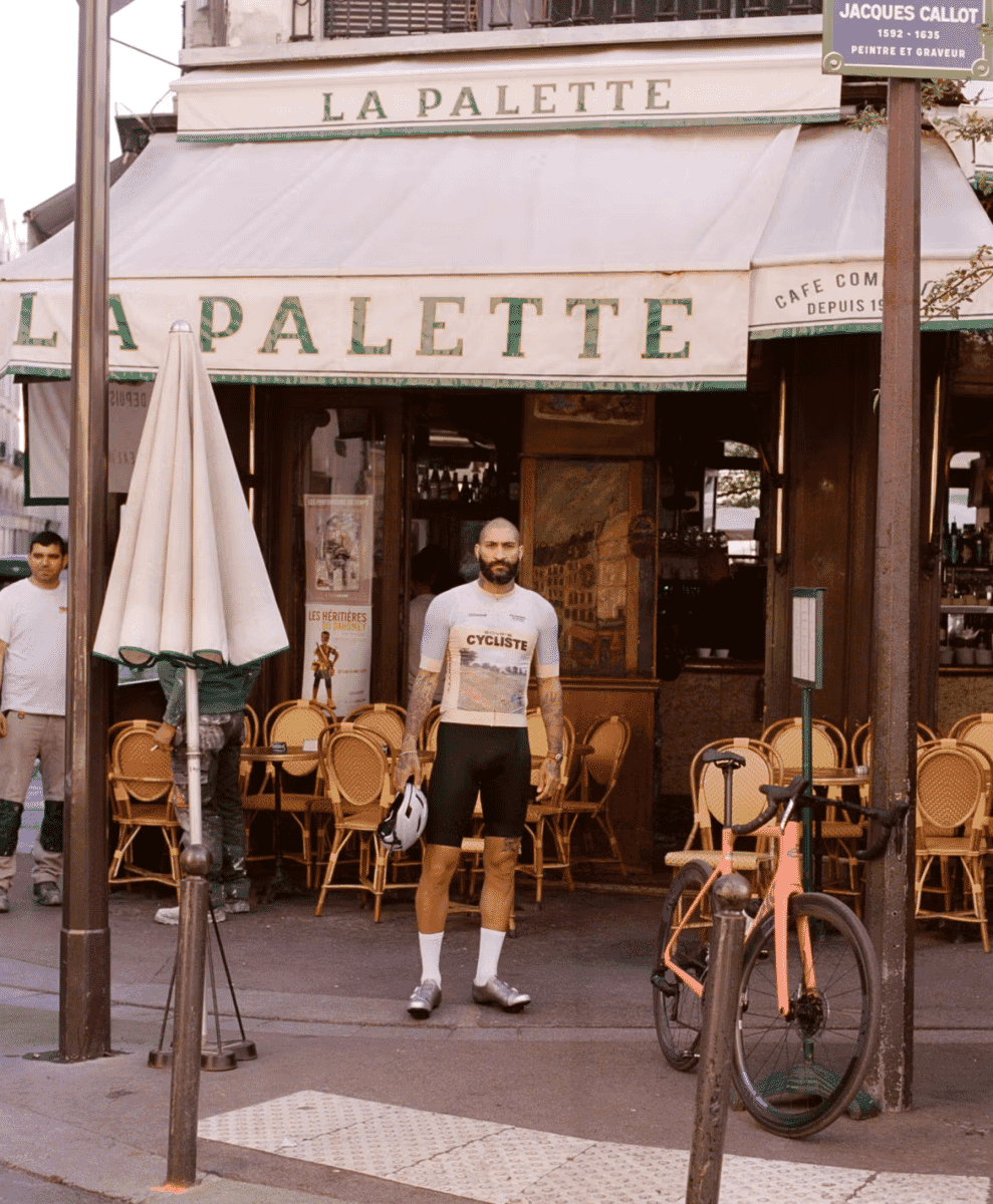 A man in a white cycling kit holding a helmet next to an orange bike outside La Palette café.