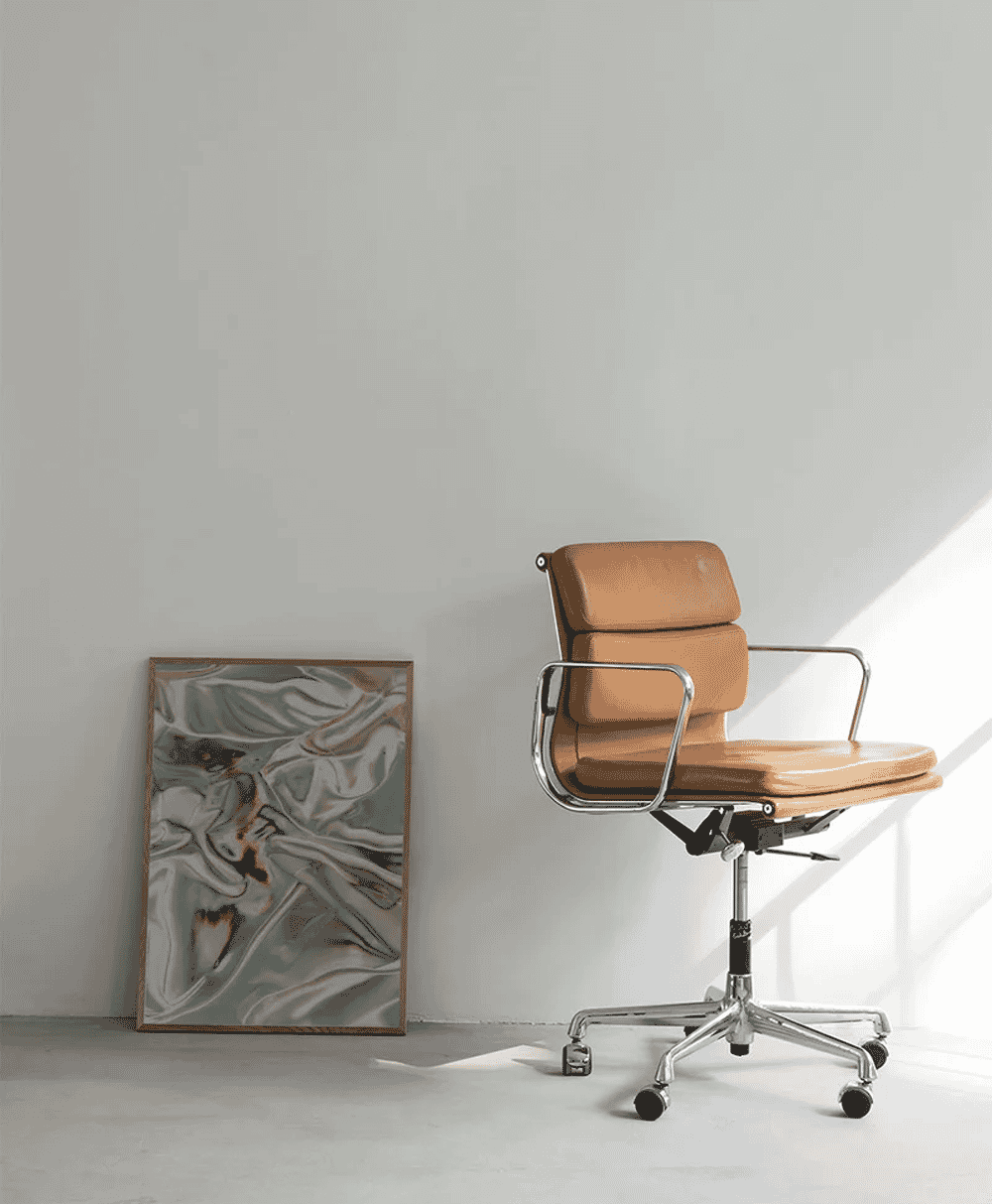 A modern tan leather office chair with a chrome frame placed next to a large abstract painting in a bright room.
