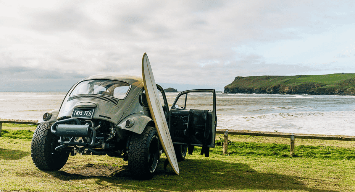 A customized VW Beetle with off-road tires and a surfboard on a grassy cliffside.