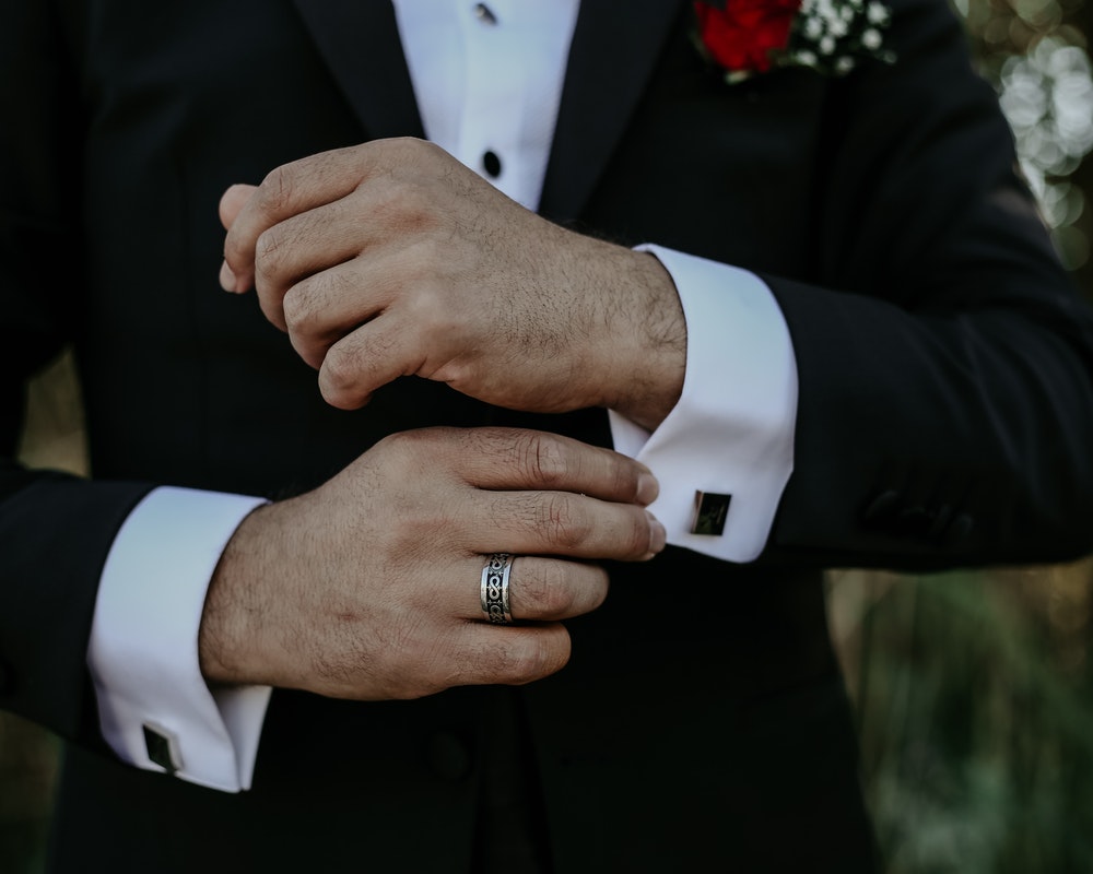 Close-up of a man in a black tuxedo adjusting white cufflinks, featuring a patterned wedding ring.