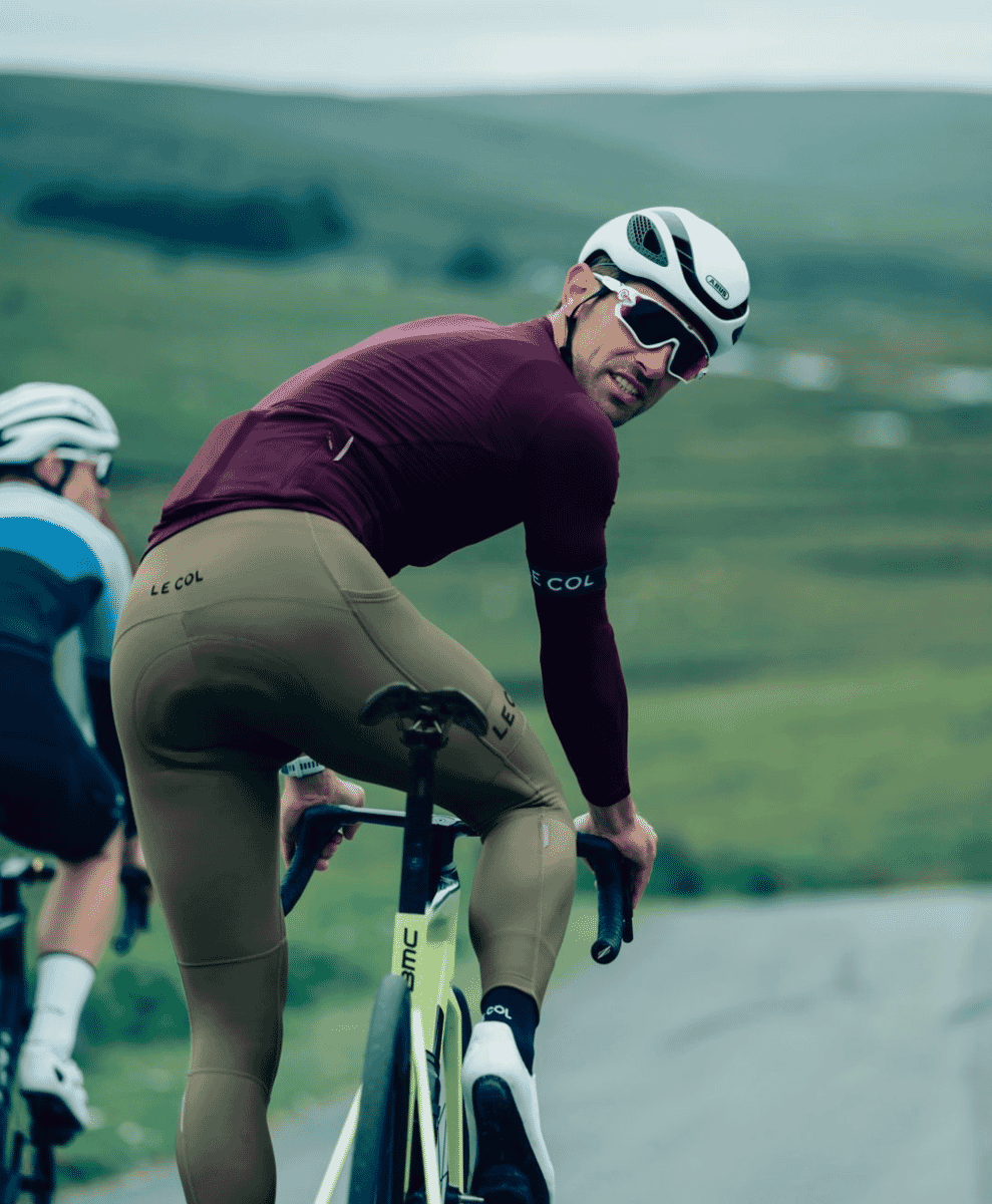 A cyclist in a maroon Rapha-style jersey and white helmet riding through a hilly rural landscape.