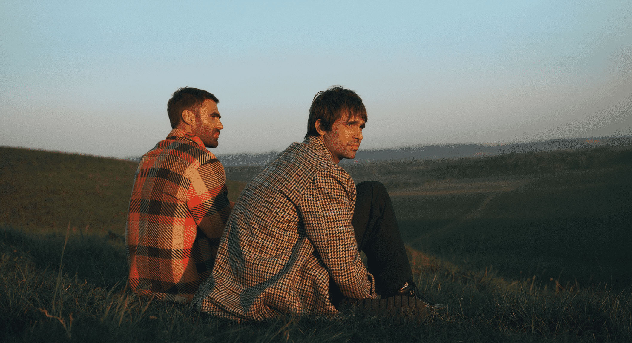 Two men sitting on a grassy hill at golden hour wearing red plaid and houndstooth coats.