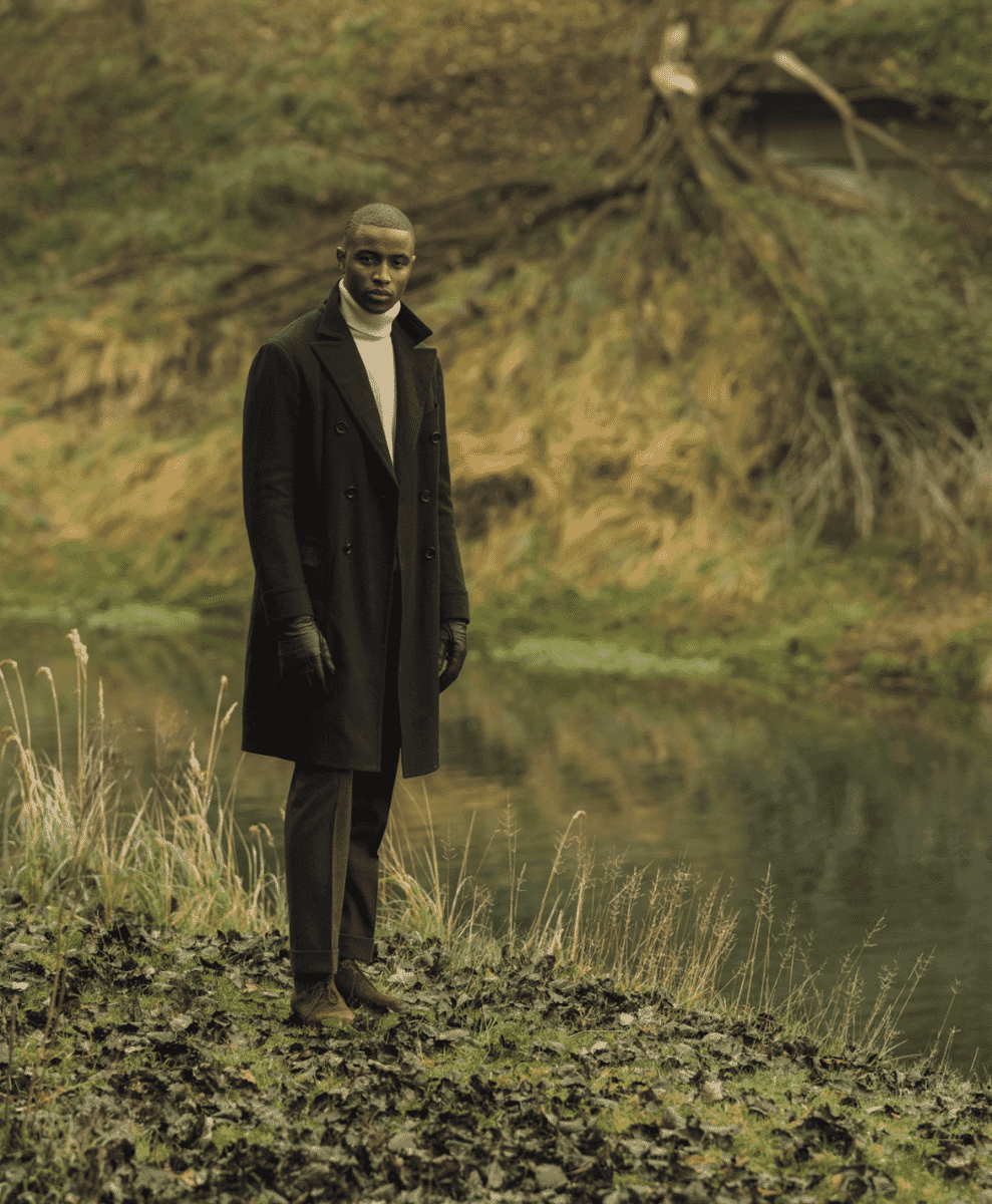 A man in a dark double-breasted coat and white turtleneck standing by a riverbank.