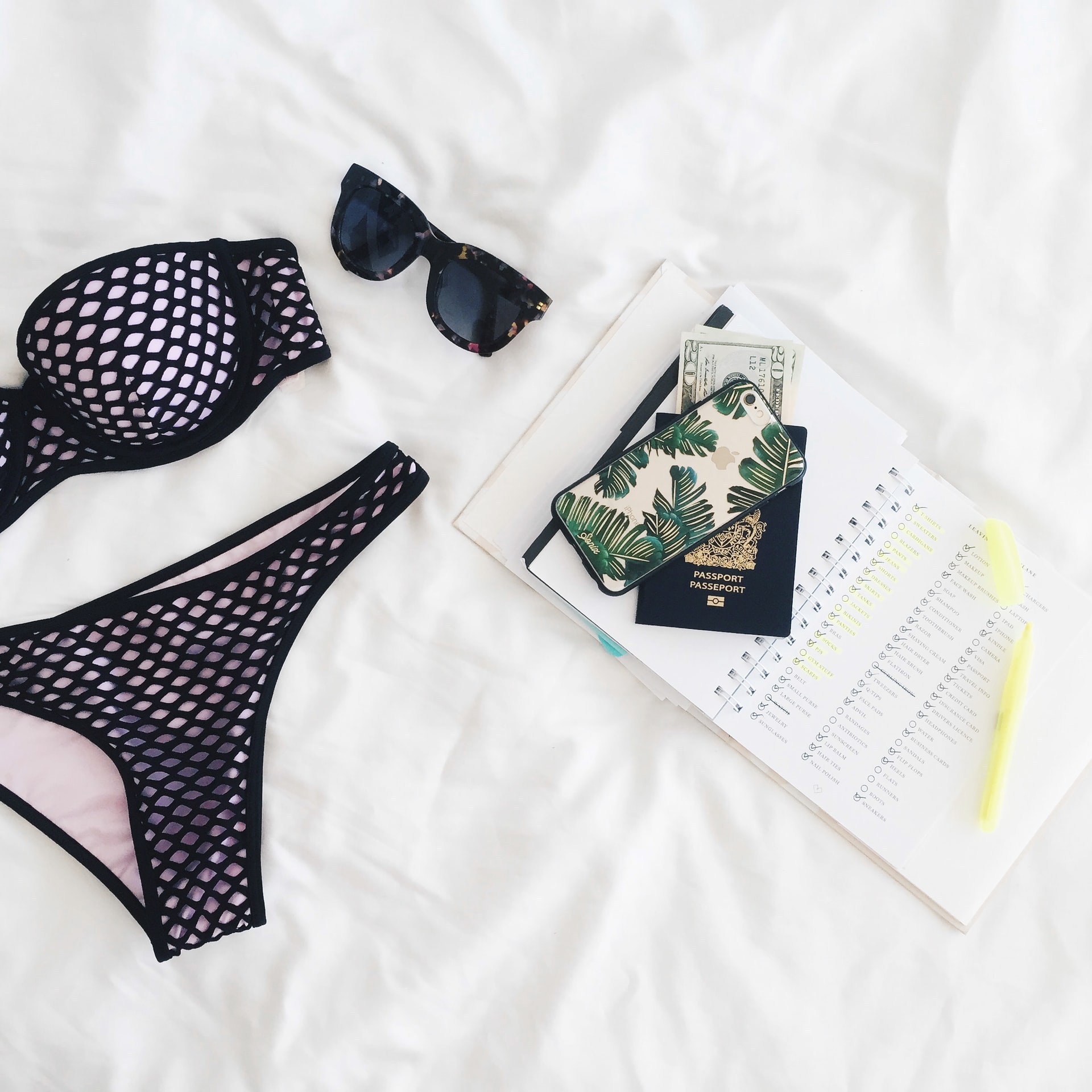 Flat lay of a black mesh bikini, tortoiseshell sunglasses, and a tropical-themed phone case on white linen.