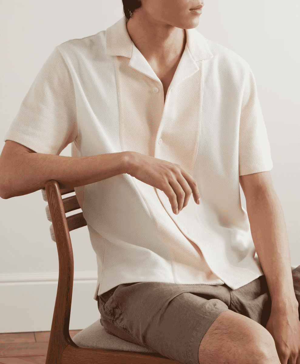 A man sitting on a wooden chair in a cream-colored knitted shirt and beige shorts.