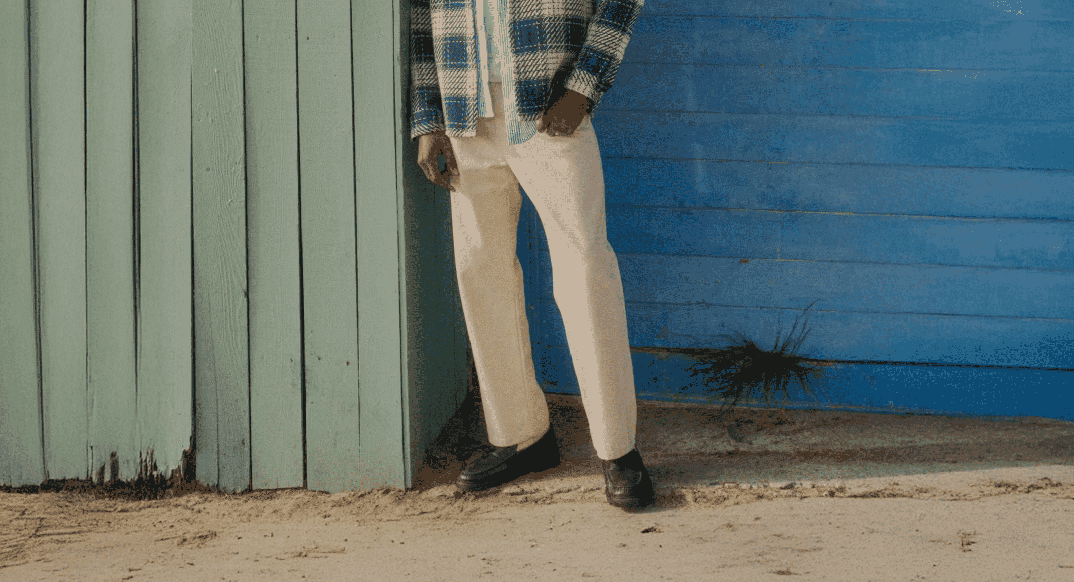 A man in a blue-and-white plaid overshirt with cream trousers standing in a sunlit outdoor setting.