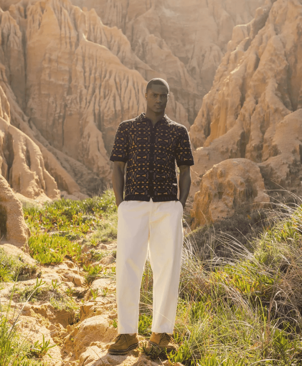 A man in a black patterned knit shirt and white wide-leg trousers in a sunlit desert canyon.