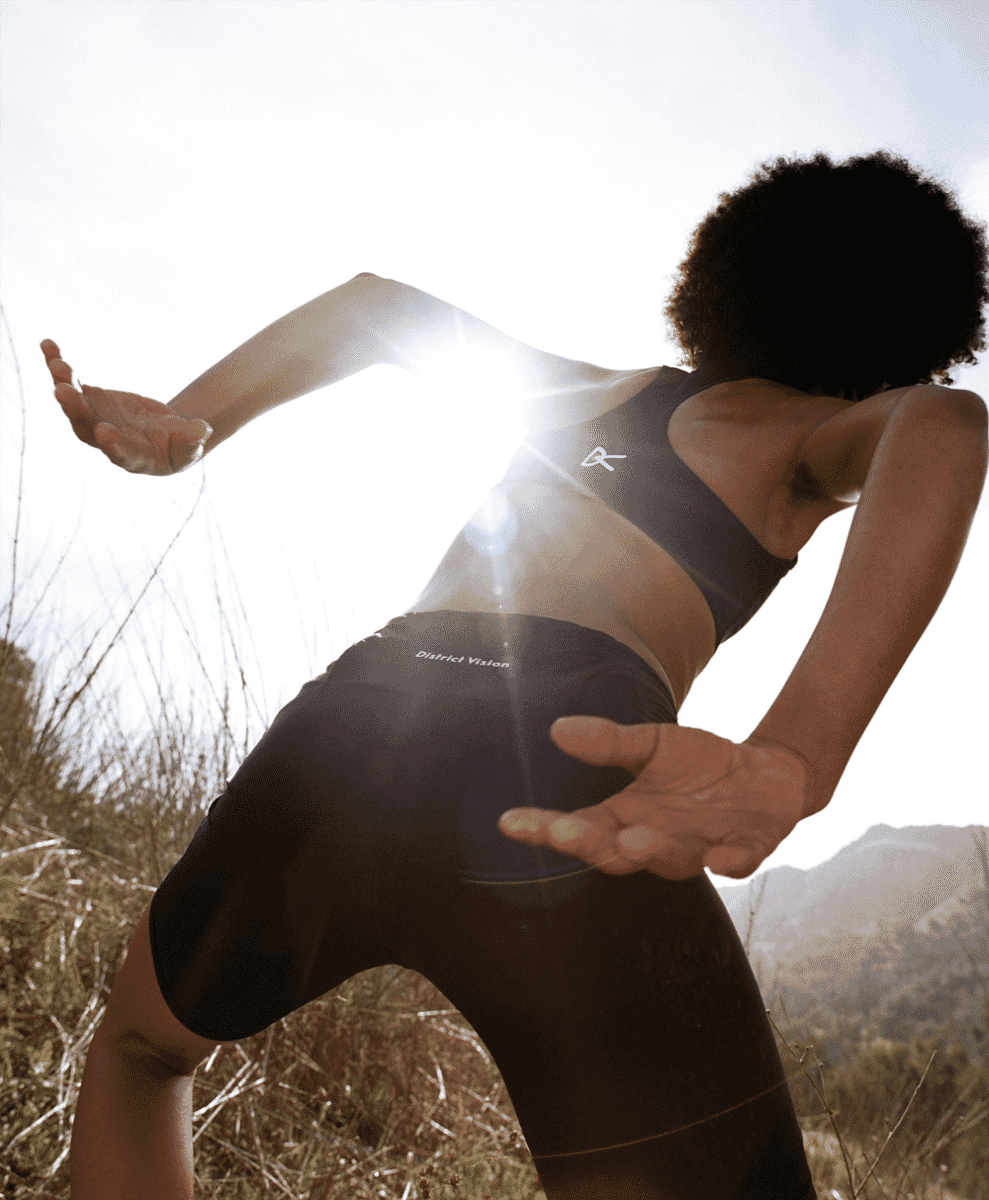 A runner in a District Vision sports bra and shorts in bright sunlight against a hilly background.