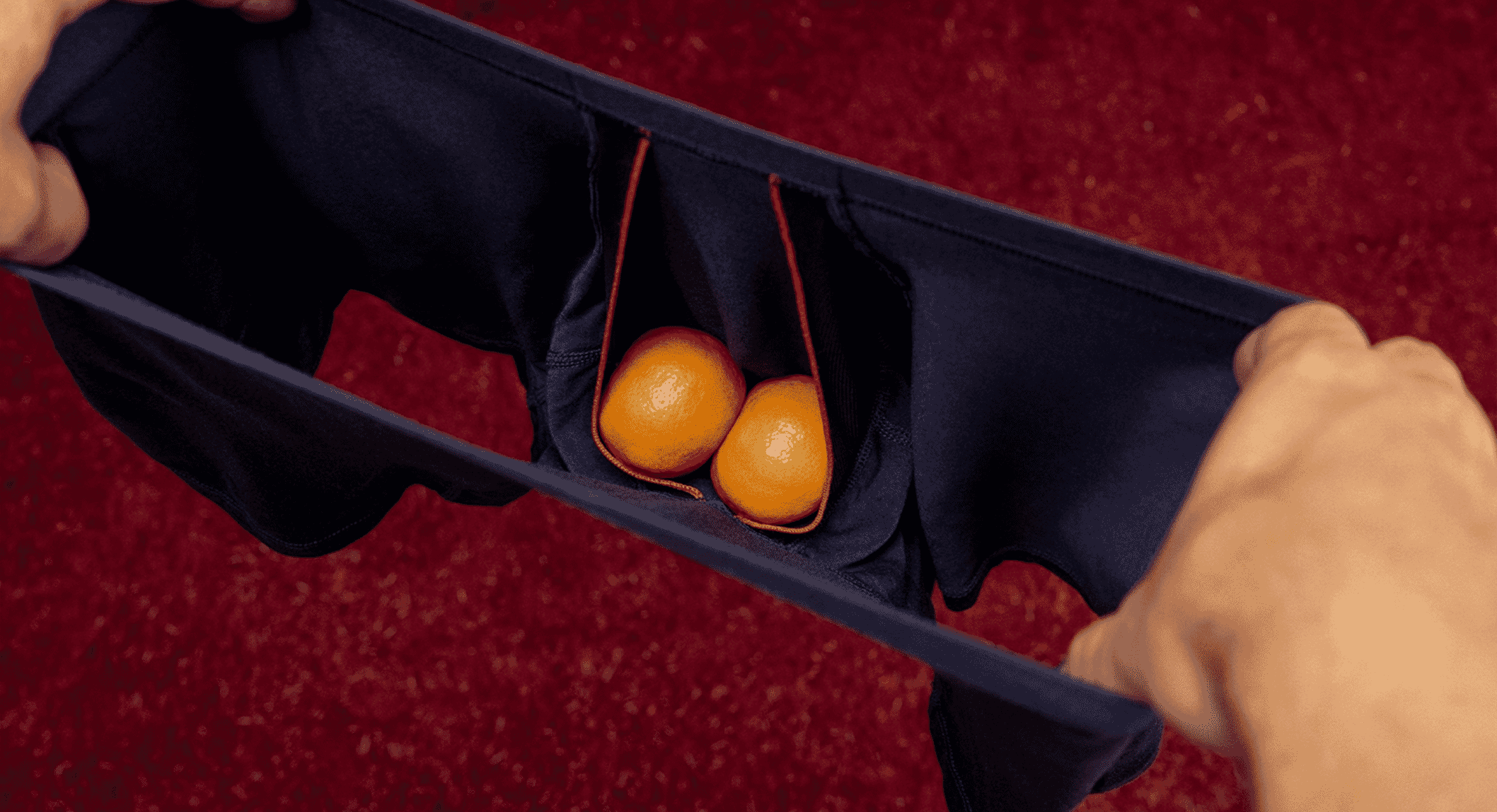 Dark-colored men's pouch underwear featuring two oranges in the support zone against a red carpet background.