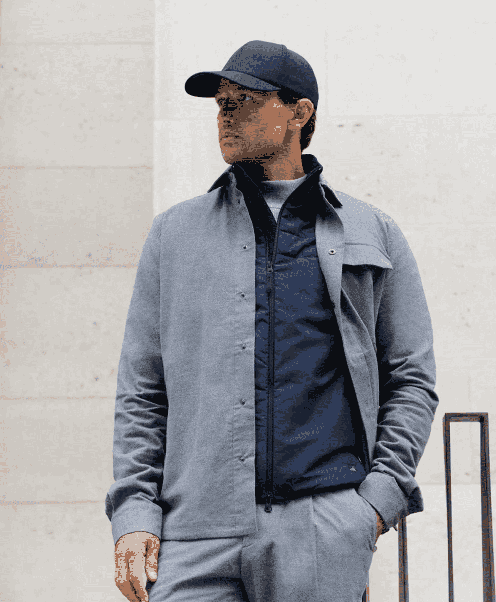 Man in a grey wool coat and dark cap standing against a concrete wall.