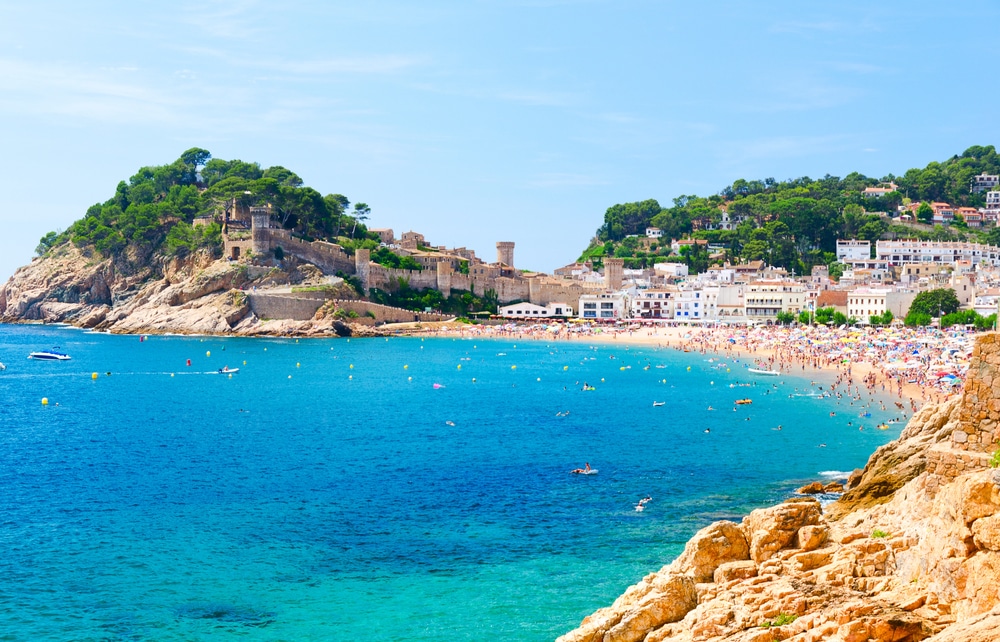 A Mediterranean beach with historic stone walls and white buildings under a bright sky.