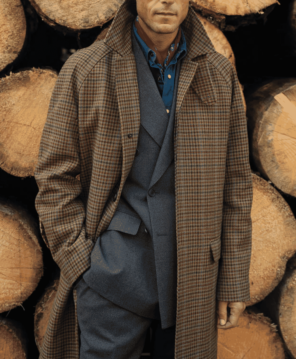 A man in a checkered wool coat standing in front of stacked wooden logs.