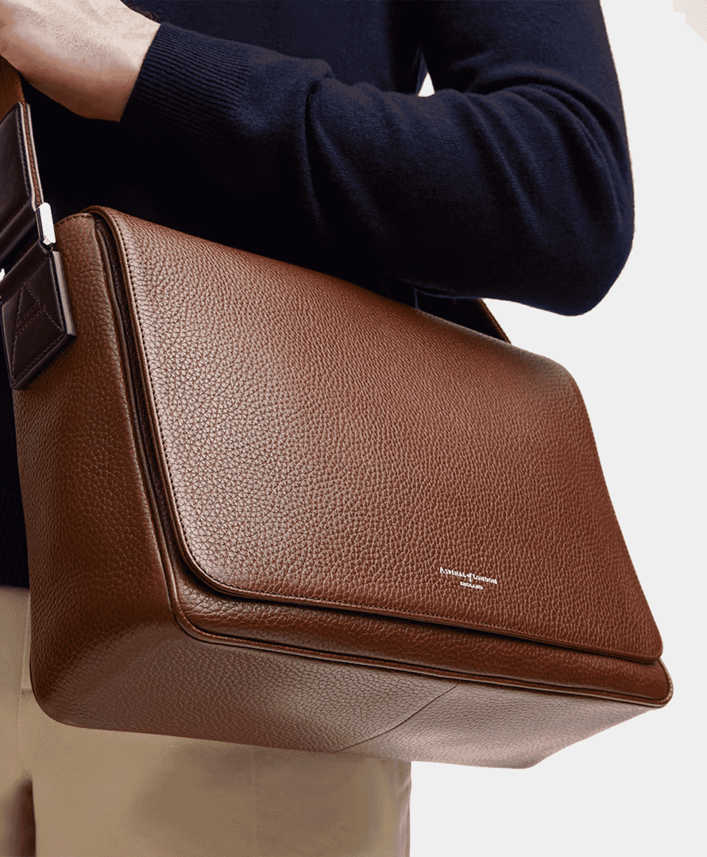 Man in a dark sweater and light trousers carrying a textured brown leather messenger bag.