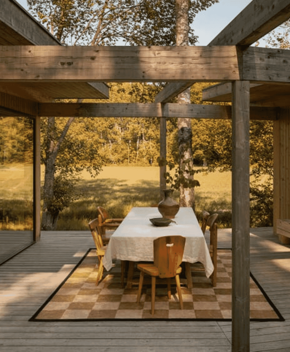 An outdoor dining set under a wooden pergola with a checkered rug set against a field backdrop.