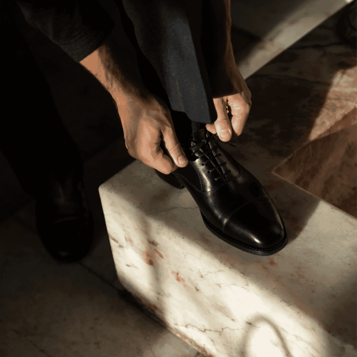 Man adjusting the laces of polished black cap-toe dress shoes while seated on a marble ledge.
