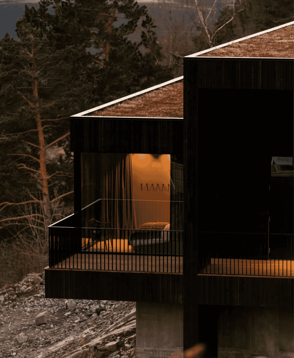 Modern architectural structure with warm interior lighting surrounded by trees and rocky terrain.