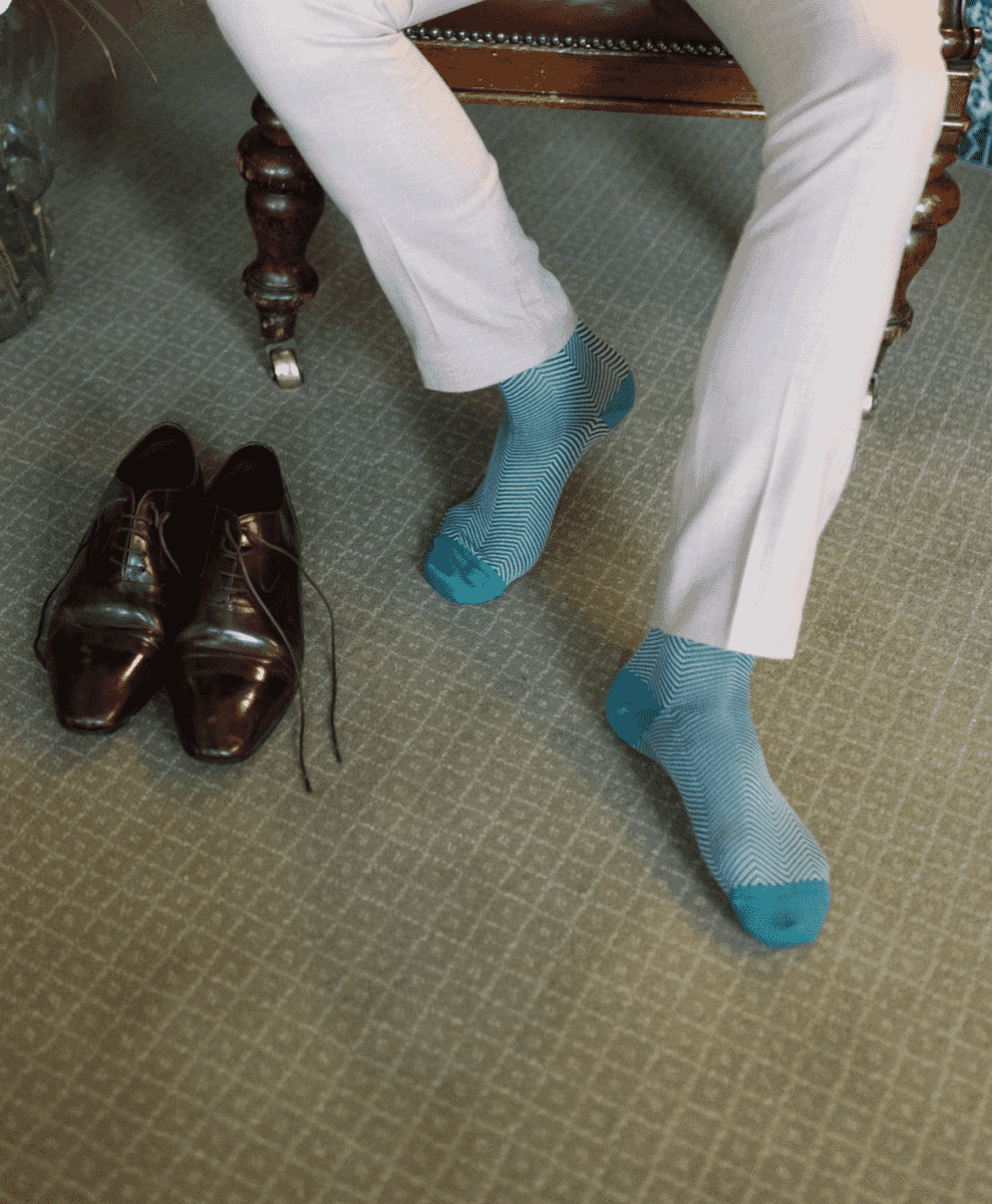 Man in white trousers and herringbone blue socks seated next to brown dress shoes.