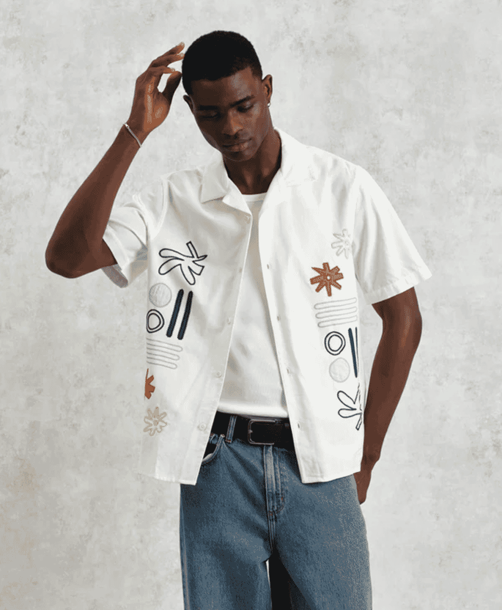 A man wearing an embroidered white camp-collar shirt layered over a white t-shirt.