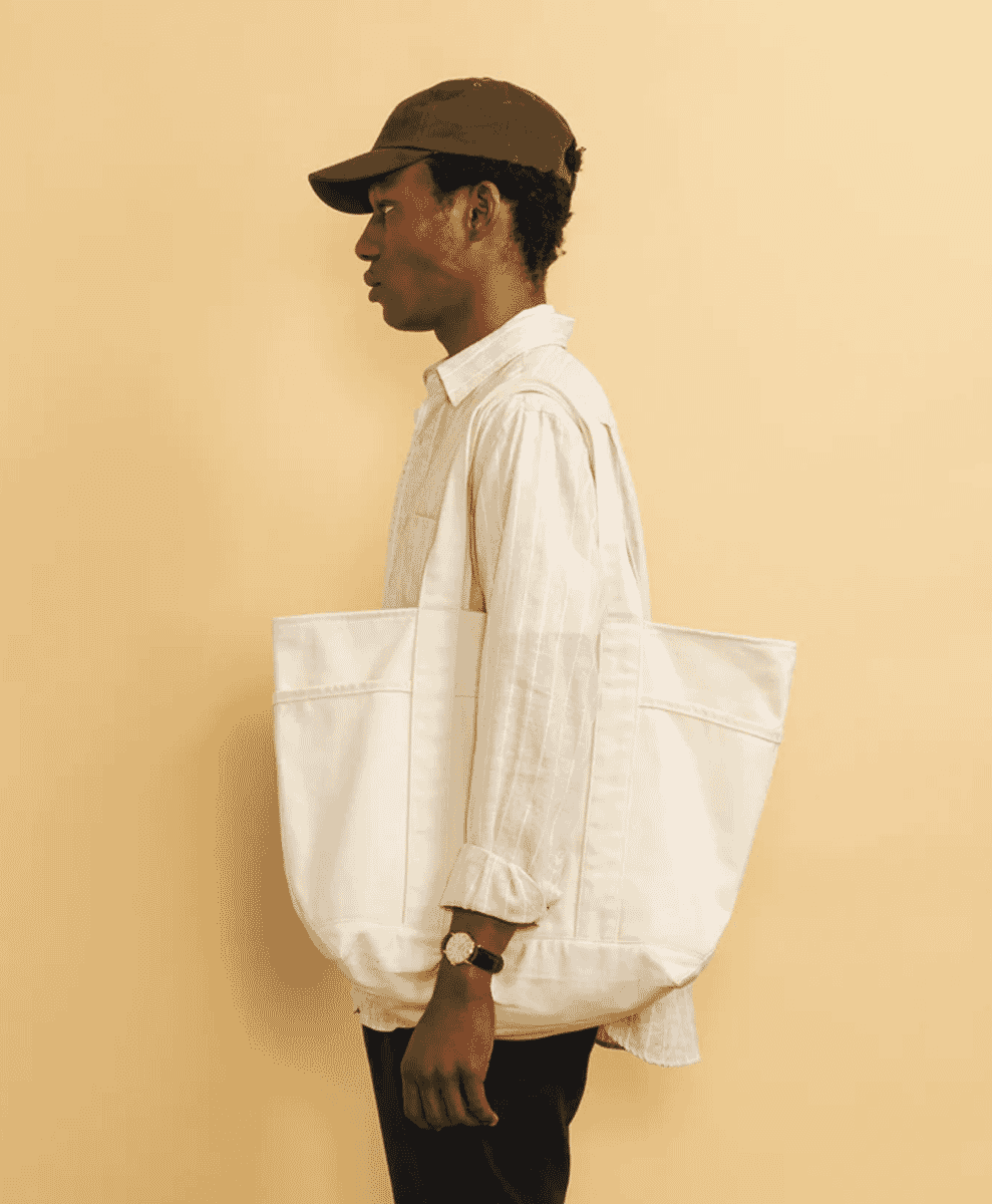 Side profile of a man in a white shirt, brown cap, and carrying a large white tote bag.