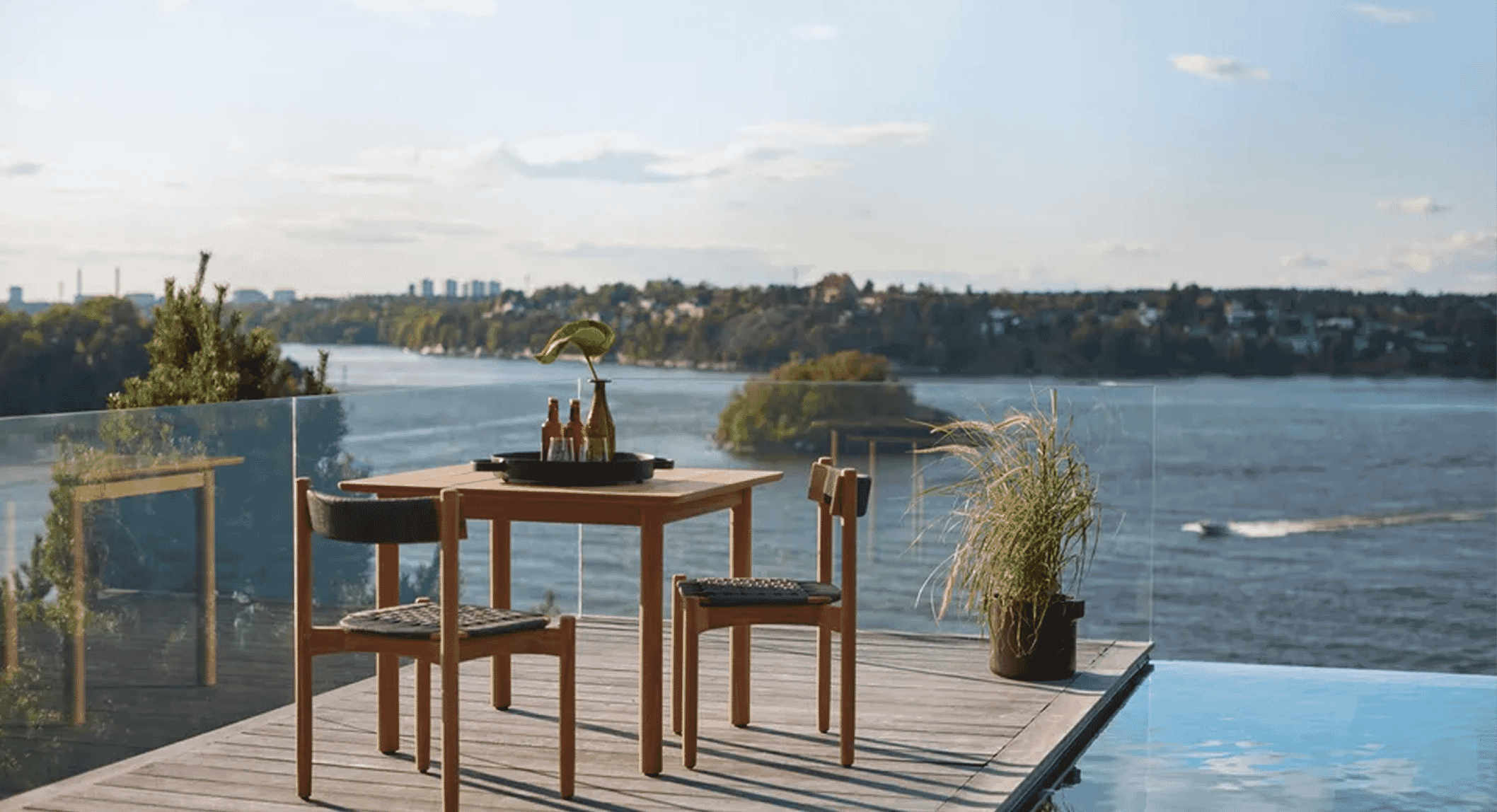 Modern wooden patio furniture overlooking a calm body of water with a city skyline in the background.