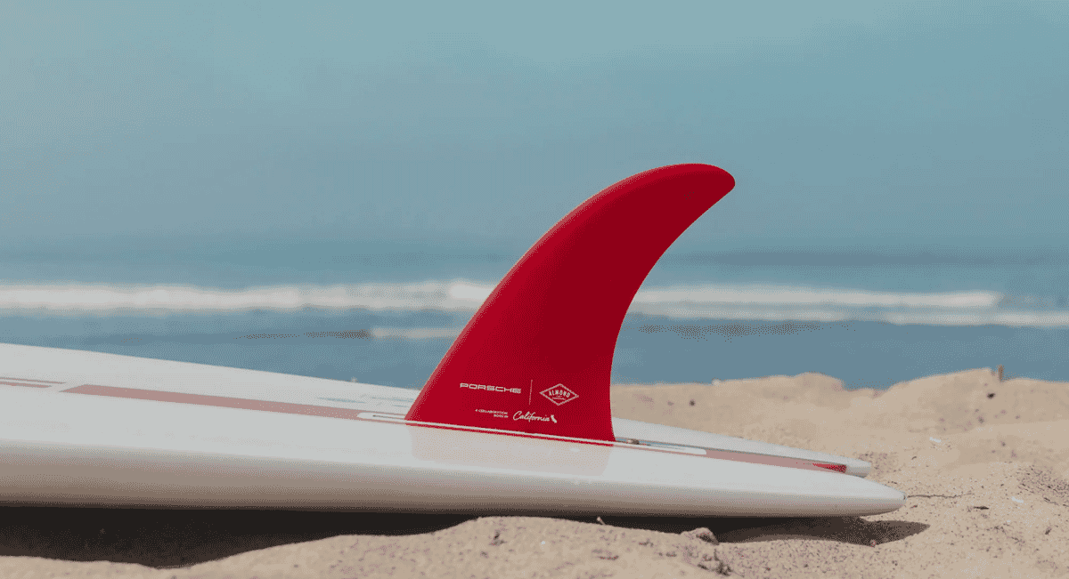 Close-up of a red Porsche-branded fin on a white surfboard resting on sand.