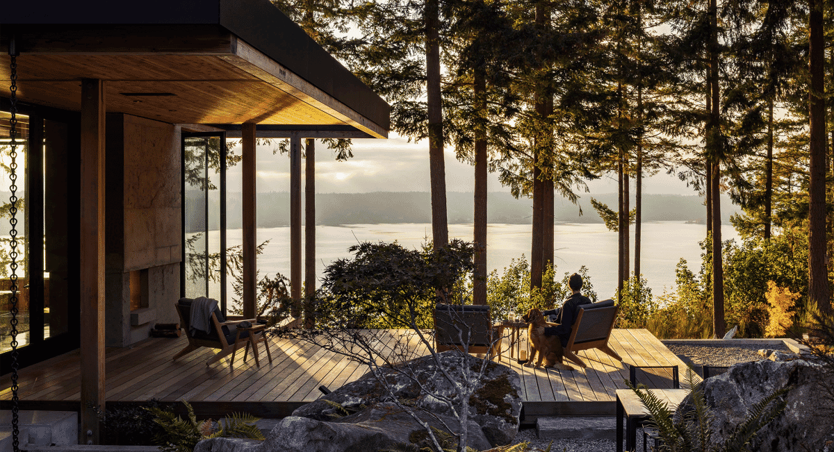 A person in relaxed dark clothing sitting on a wooden deck with a dog overlooking the water.
