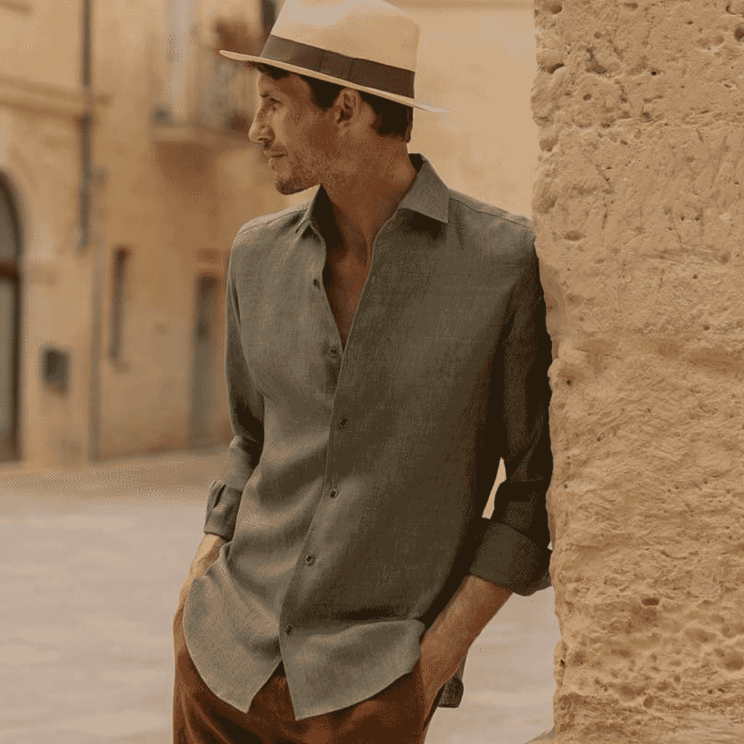Man in a gray linen button-up shirt and brown trousers wearing a cream fedora on a sun-drenched European street.
