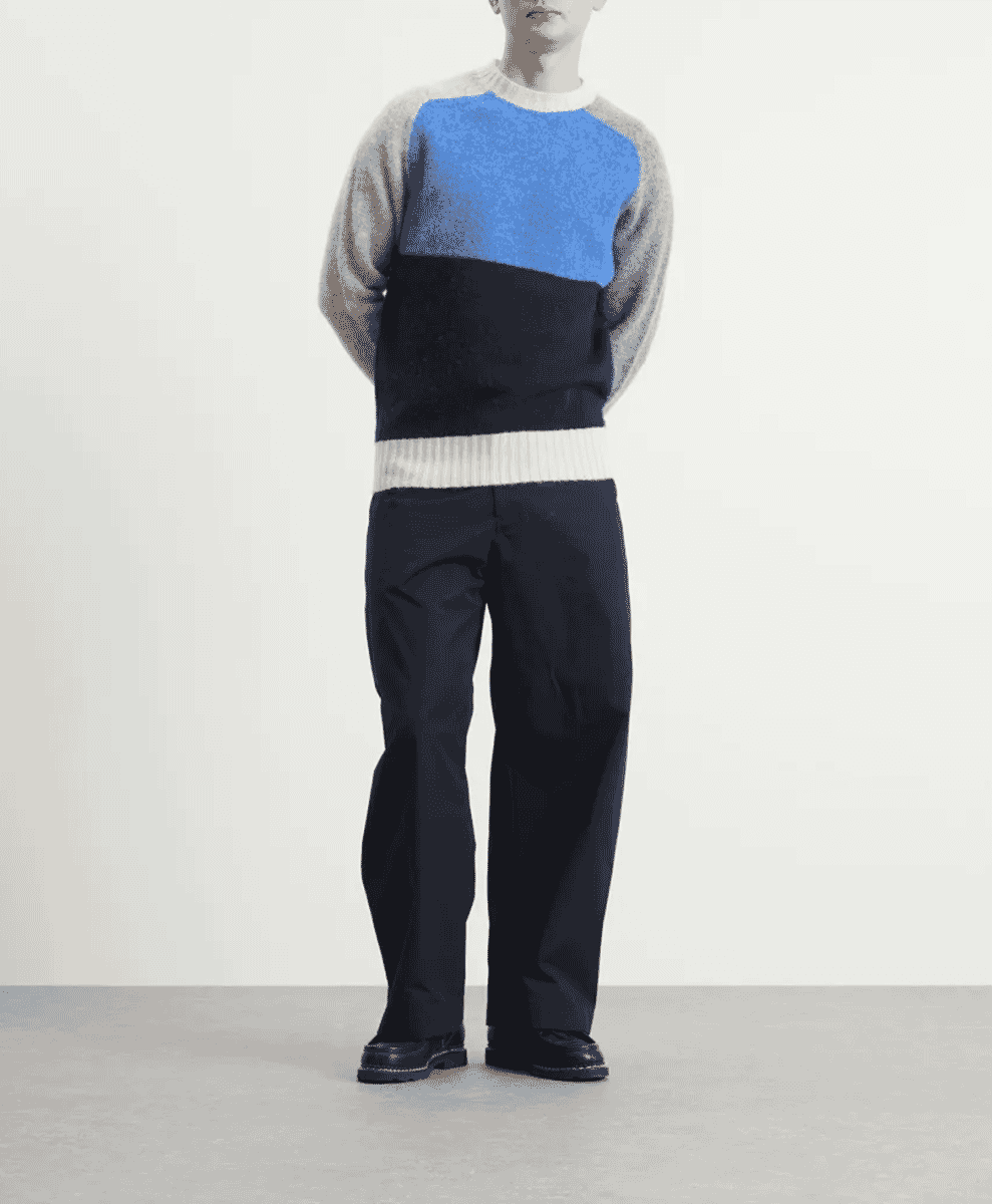 A model wearing a blue, navy, and grey color-block knit sweater with dark trousers and black shoes.