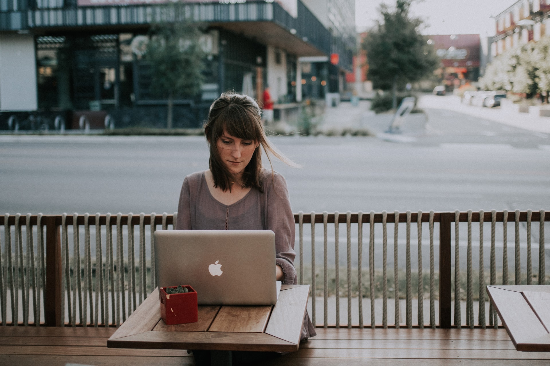 A woman in a muted mauve V-neck top using a MacBook at a wooden table in an outdoor setting.