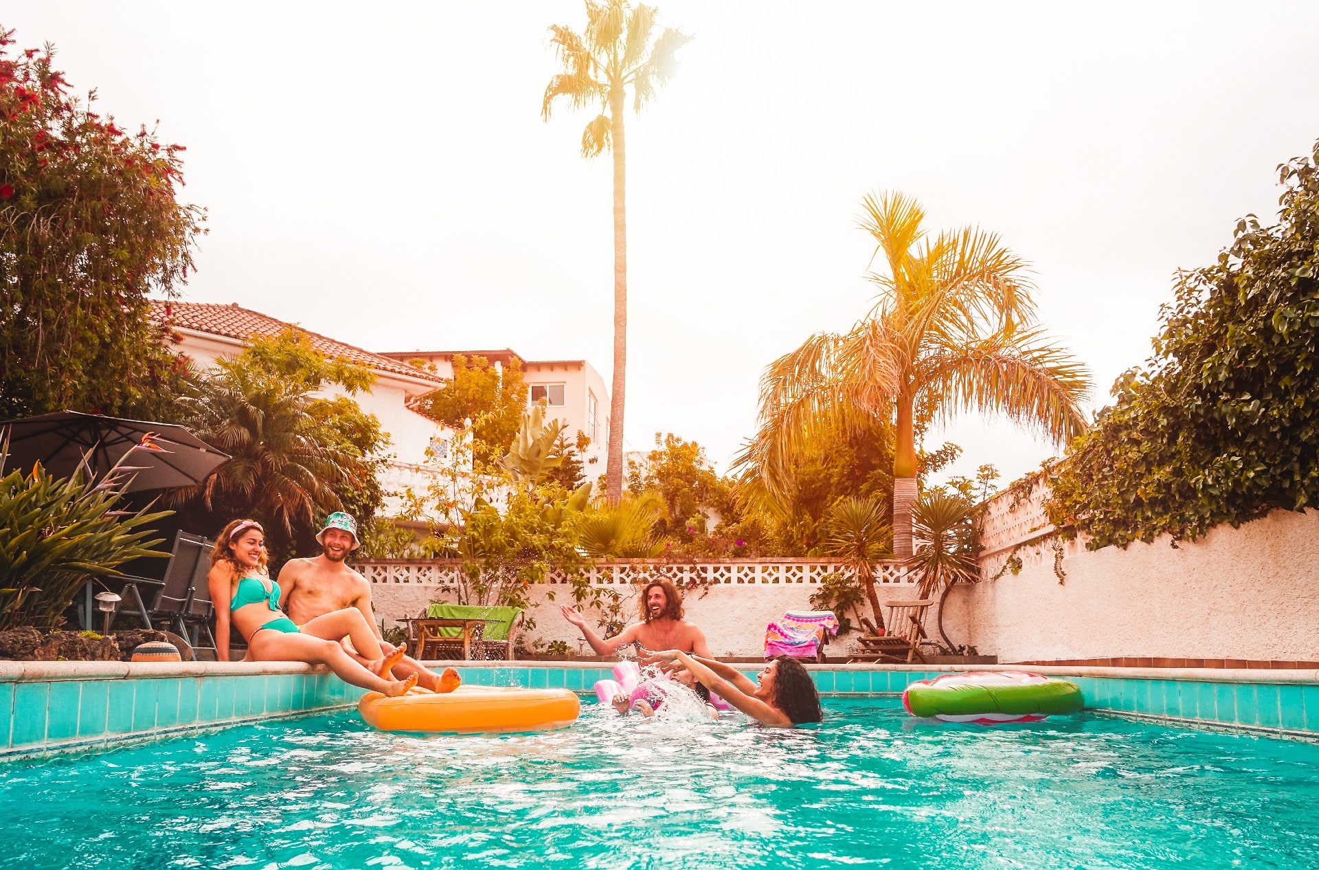 Friends in designer swimwear and casual beachwear enjoying a sunny pool party with palm trees and tropical plants.